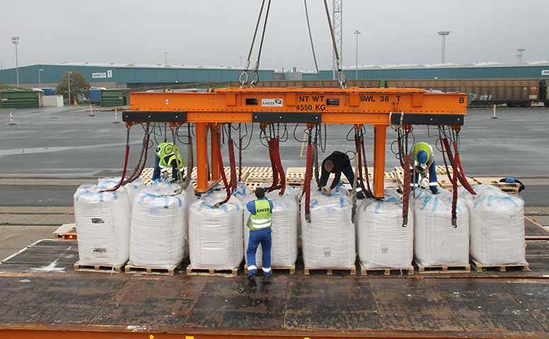 Port Specific Services: People in yellow Anker Schiffahrt safety vests are unloading big bags with orange specialized equipment. In the background, a large warehouse can be seen
