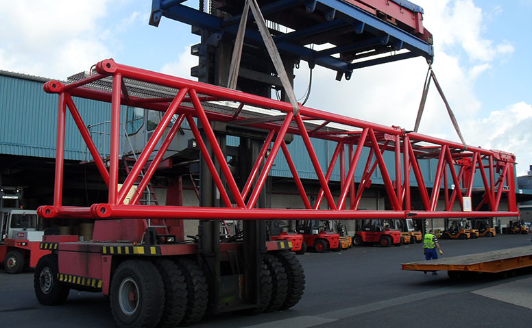 Port Specific Services: A reach stacker, under the supervision of a person in a yellow anchor safety vest, is lifting a break-bulk cargo onto a truck tractor. Forklifts and a large warehouse can be seen in the background 