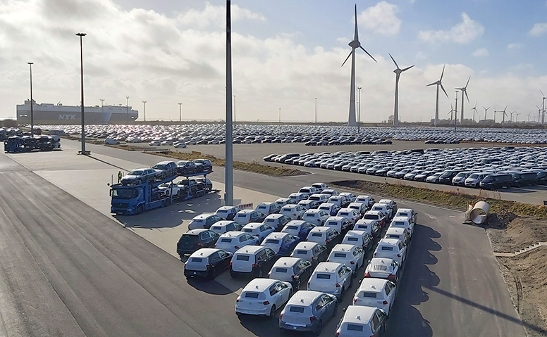 Automobile handling: Photograph of the automobile handling area of Anker Schiffahrt in Emden, showing cars wrapped in white protective film, truck car transporters, wind turbines, and a large car carrier in the background