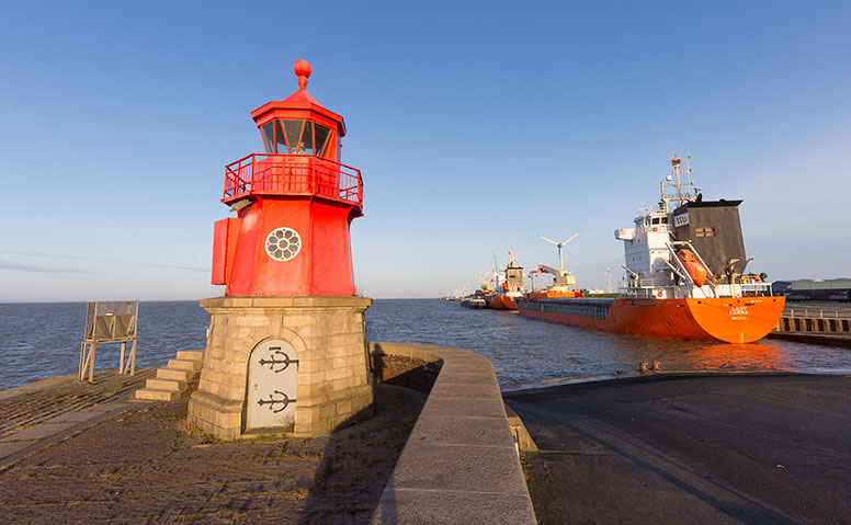 Port Specific Services: The famous red Emder lighthouse stands at the pier on a sunny day with blue skies. To the right, orange cargo ships are docked at the wharf