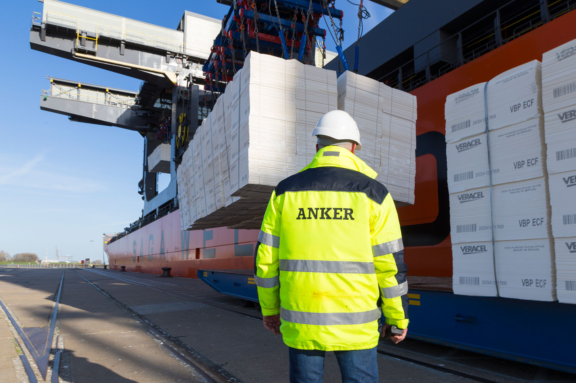 Handling of Forest Products: A crane loading forest products from a freighter onto a tractor, under the supervision of a person in a yellow Anker Schiffahrt warning jacket