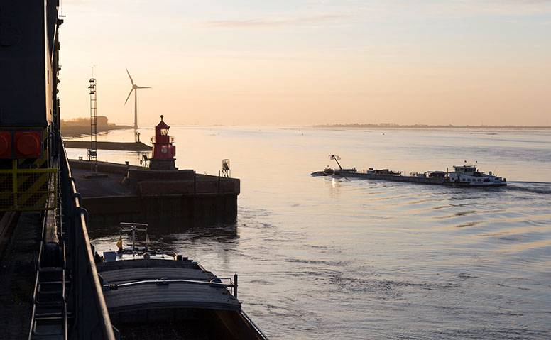 Ships Agency: Der Emder Hafen beim Sonnenaufgang. Zu sehen sind ein Frachtschiff und der Emder Leuchtturm auf der Mole. Im hintergrund ist ein Windrad