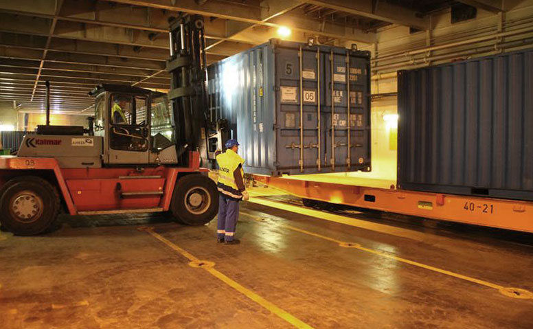 Port Specific Services: Under the supervision of a person in a yellow safety vest, a forklift transports a blue container onto a tractor inside a warehouse