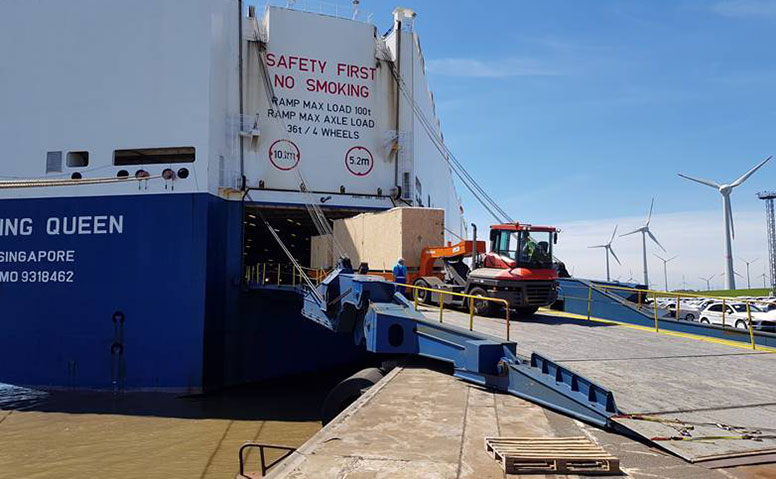 Port Specific Services: A red tractor is loading bulky goods onto a large cargo ship. Wind turbines can be seen in the background