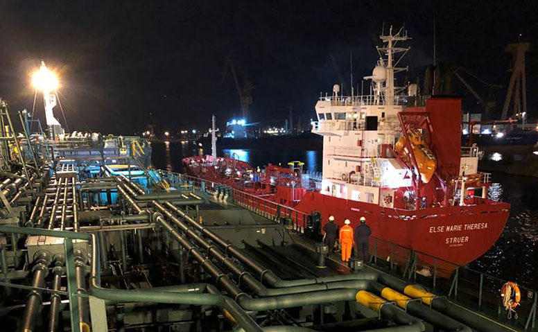 Ships Agency: Emder Hafen bei Nacht. 3 Personen in Arbeitskleidung befinden sich auf einem Tanker und schauen auf ein anlegendes rotes Frachtschiff